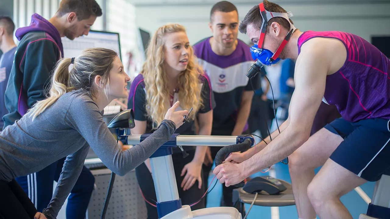 Five sport science students carrying out research in a lab. One person is wearing an oxygen mask whilst cycling on an exercise bike, another person is holding a stopwatch.