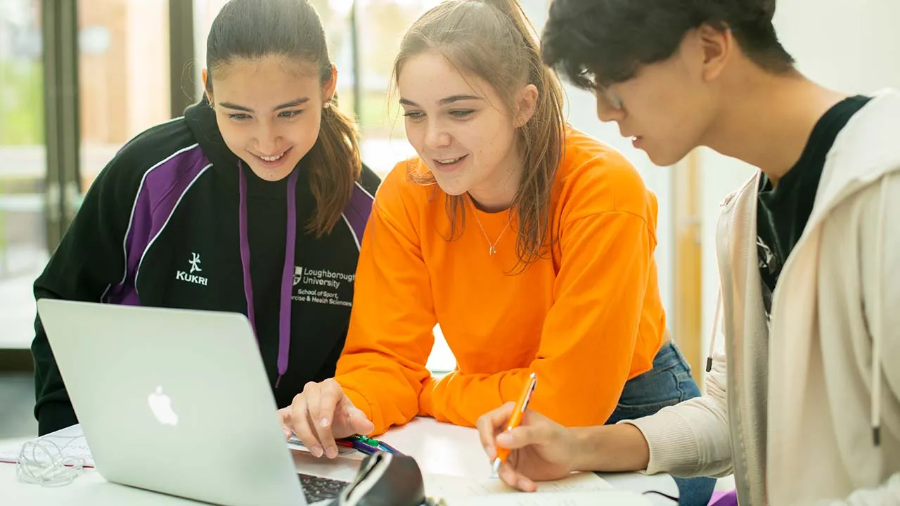 Three people stood round an open laptop looking at what is on the screen and taking notes