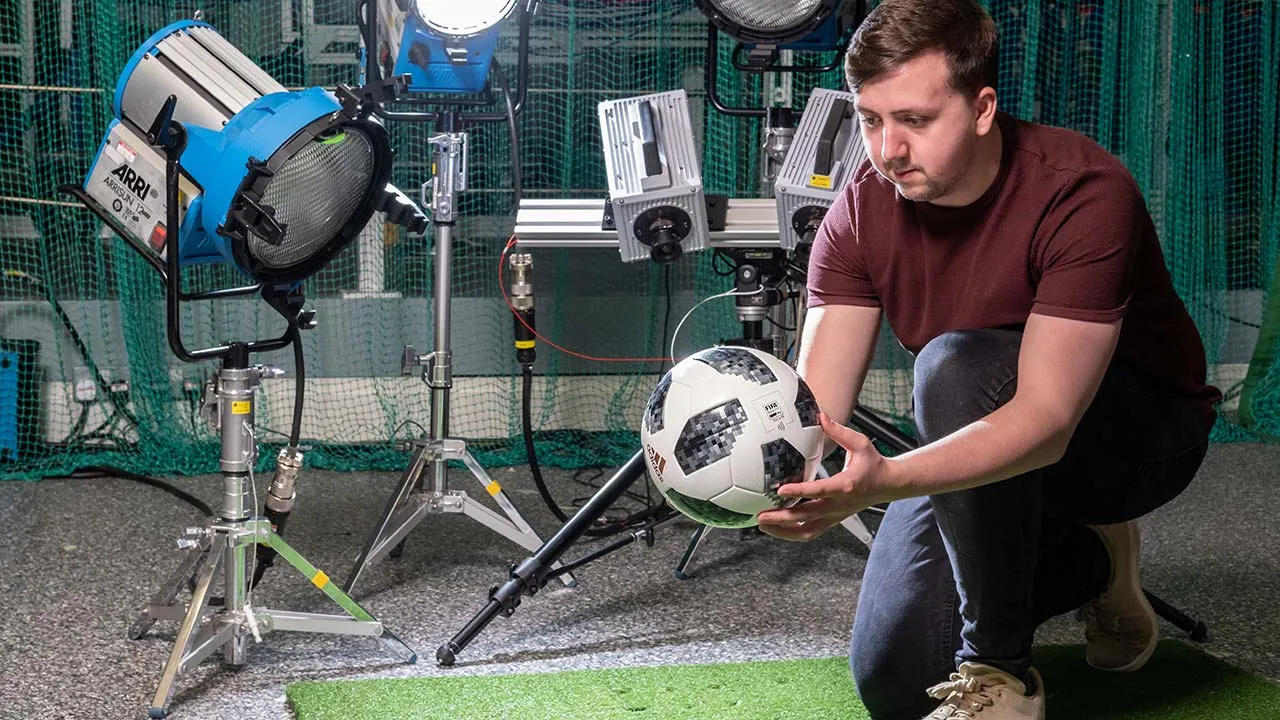 Person positioning a football in a sports technology laboratory surrounded by professional lighting equipment, high-speed cameras, and measurement instruments.