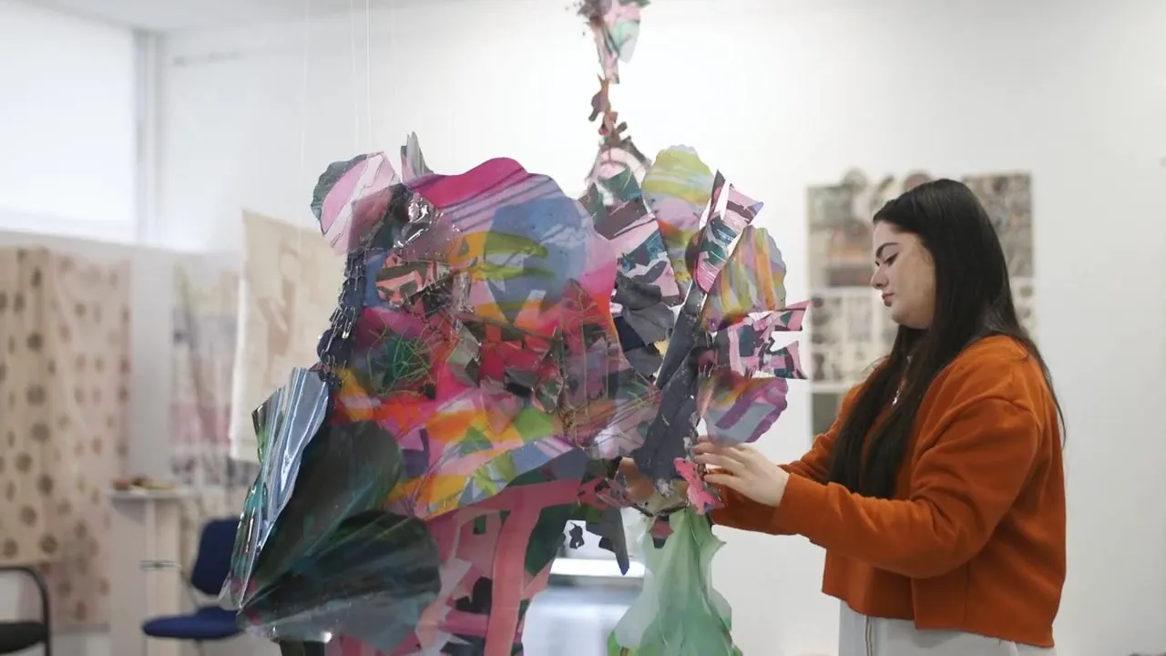 A woman is focused on creating a large artwork, surrounded by paint and brushes in a bright studio.