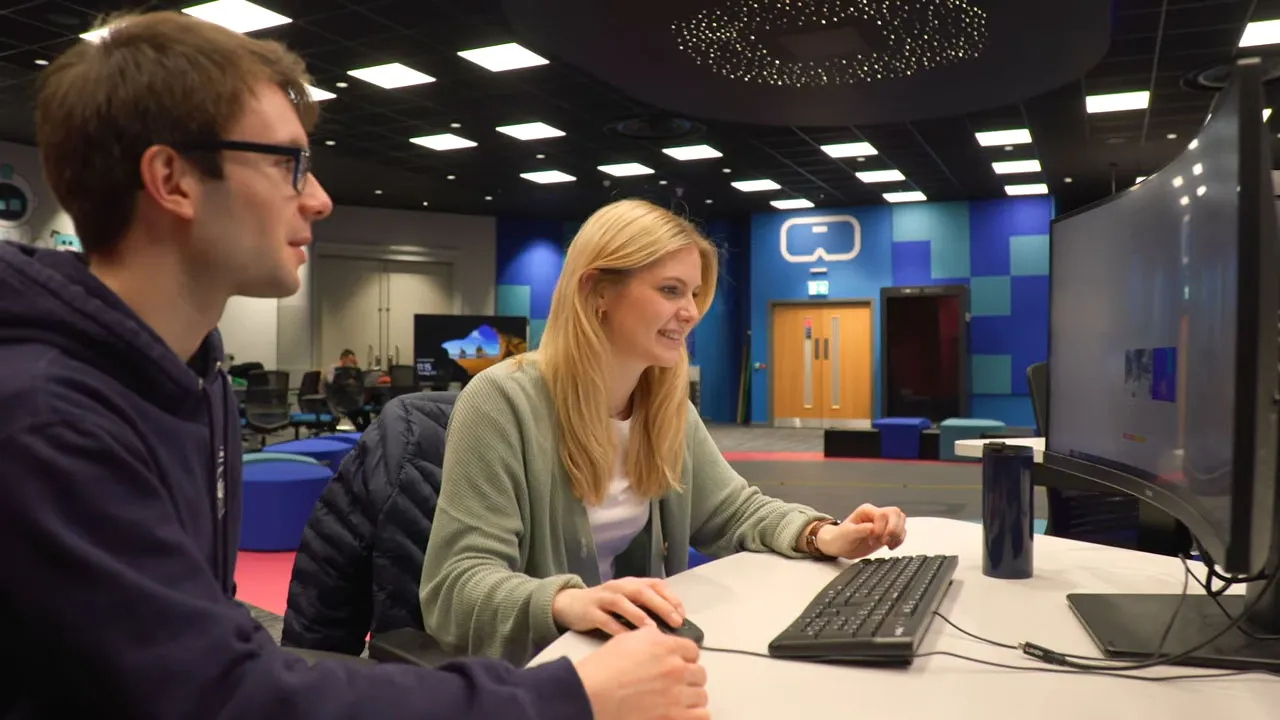 Two people sit at a desk in a modern office, looking at a curved computer monitor. One person is using a mouse and keyboard while the other observes.