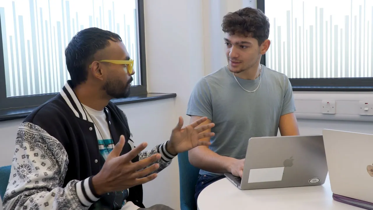Two students sat at a table talking. One has a laptop on the desk in front of them