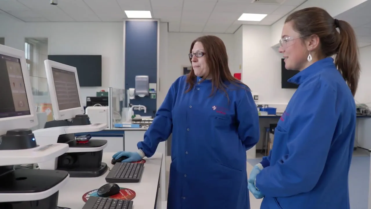 Two people wearing blue lab coats standing at a desk looking at computer screens