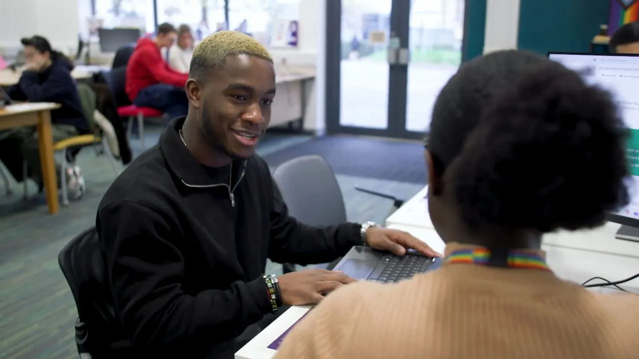 A male student wearing a black jumper sat talking to a lecturer at a desk in a study space