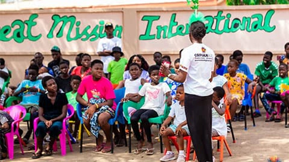 A group of children and young adults in Ghana. They are all pointing towards and listening to someone who is speaking in a white t-shirt that reads Orphan Art.