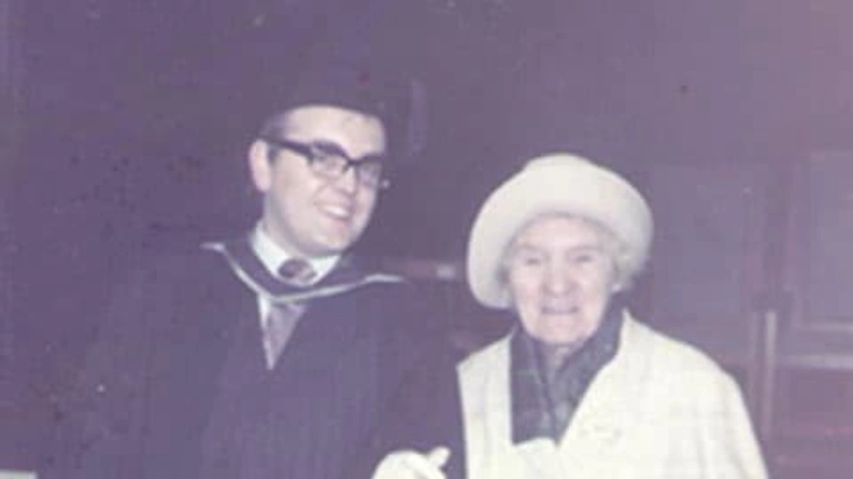 Barry in his graduation gown and hat with his grandmother. They are smiling together.