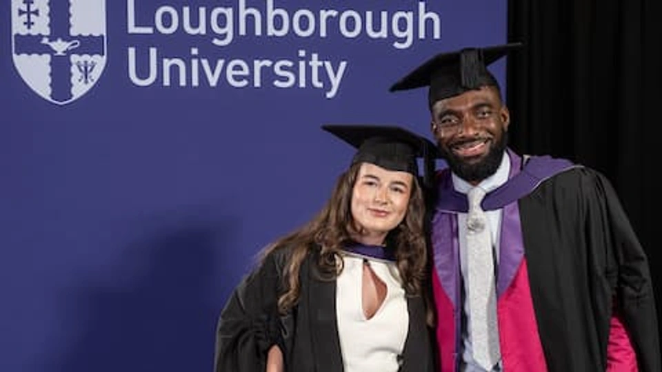 Libby and Jaivani wearing graduation attire. They stand in front of an African Violet backdrop that has a Loughborough University logo on.