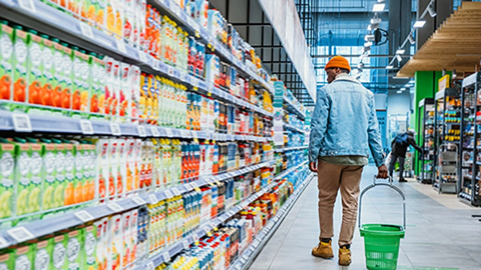 Person with green plastic basket walks looking at shelves with different goods in modern supermarket department