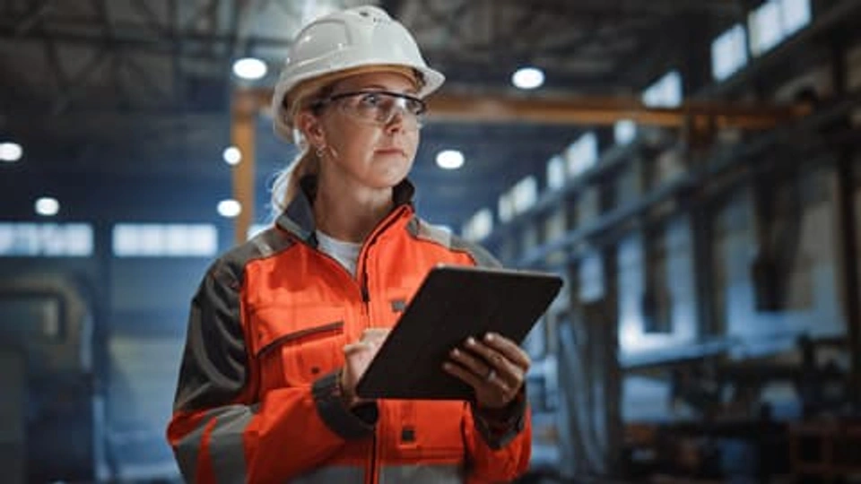 A worker in an industrial building, wearing a hard hat, hi vis clothing, and holding a tablet computer.