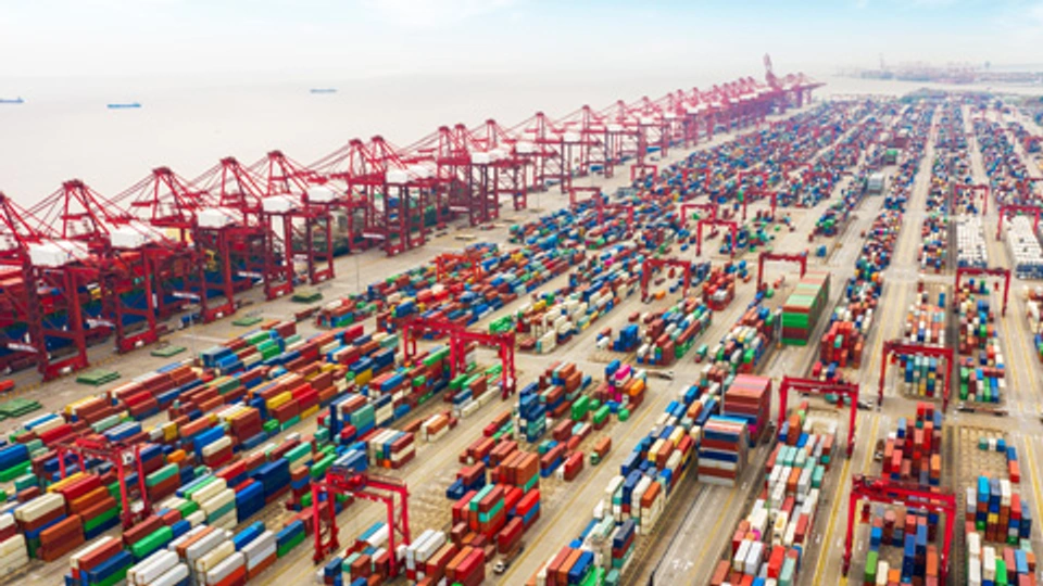 Aerial photo of a busy shipping port with rows of multicolored cargo containers, red cranes, and cargo ships docked in the harbor depicting global maritime logistics.