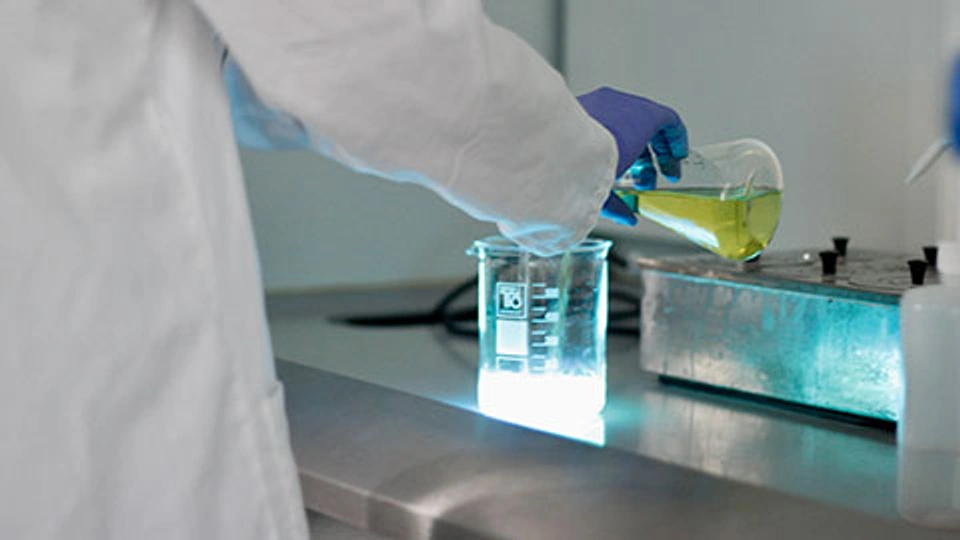 A person in a lab coat and gloves pouring a liquid from glassware