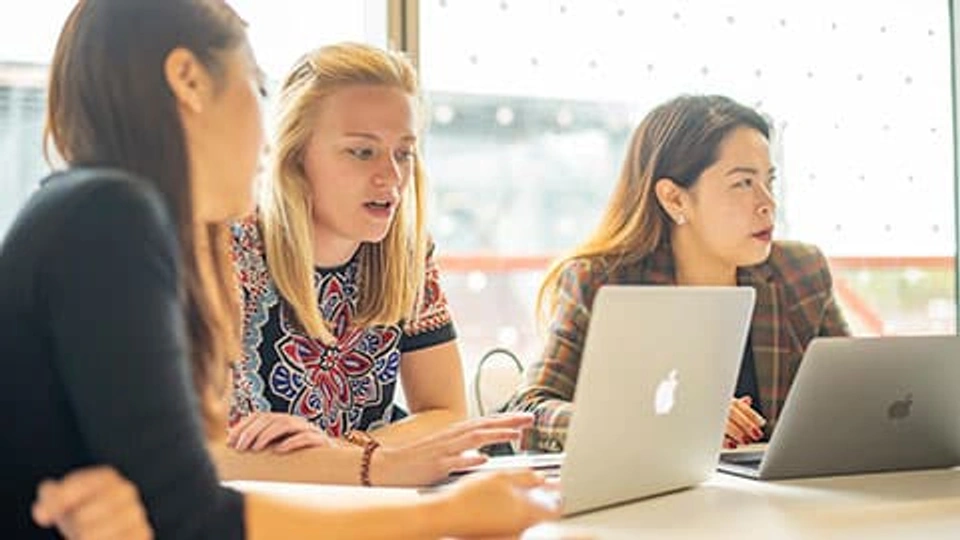 Three girls sat at a table with two of them looking at a laptop screen