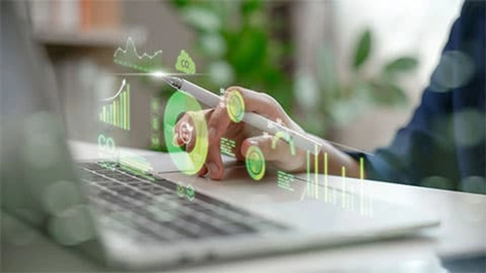 A person holds a digital pen while interacting with green, glowing sustainability charts and carbon footprint data projected over a laptop keyboard.