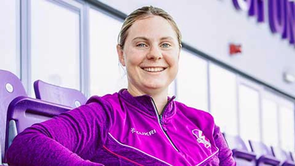 Katie Sorenson wearing a Lightning quarter zip, sitting in the stand at the stadium, smiling whilst looking directly at the camera