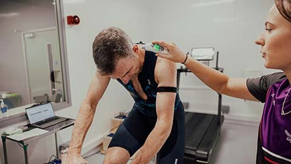 A client is on a bike doing a session, while a physiologist holds a thermometer in his ear.