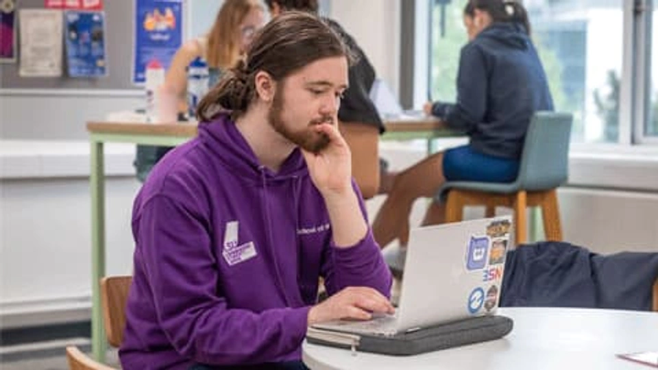 A male student wearing a purple hoody working on his laptop