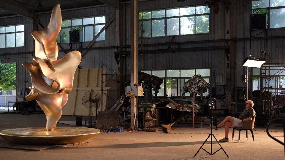 A large abstract metallic sculpture stands on a circular base inside an industrial workshop, lit by warm studio lights, while artist John Atkin sits on a chair to the right facing it, with photography equipment, windows, and workshop tools visible in the background.