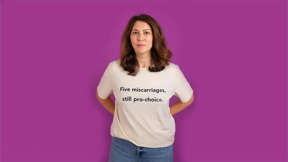 A woman wearing a t-shirt against a plain background which says five miscarriages, still pro-choice
