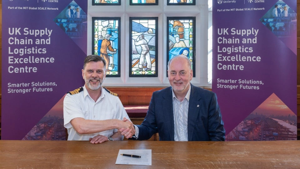 Two men sat at a table shaking hands looking into the camera
