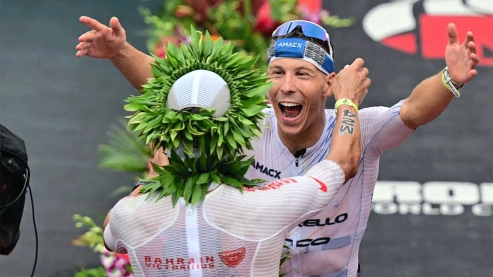 Two triathlon athletes in racing suits embrace at the finish area after a competition, with one athlete wearing a leafy victory wreath and flowers visible in the background.