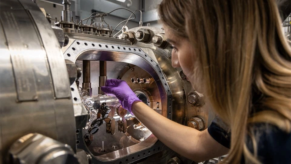 A woman wearing purple gloves carefully adjusts a transparent glass component inside a large stainless-steel scientific vacuum chamber filled with metal fittings, bolts, and precision instruments.