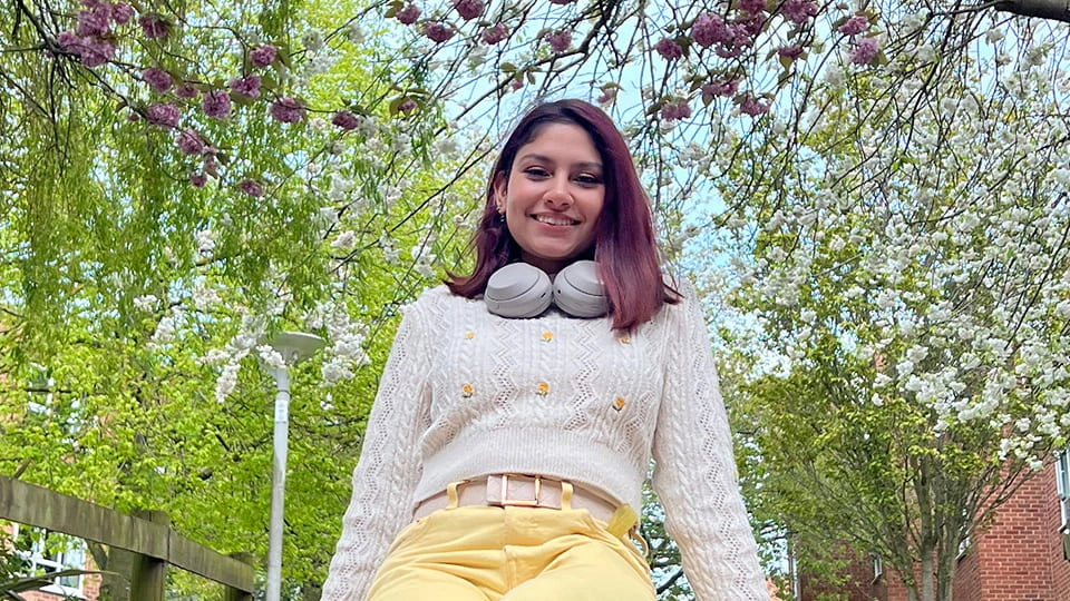 a female student sitting outside on a spring day smiling toward the camera, there are many trees surrounding her with white and purple blossom.