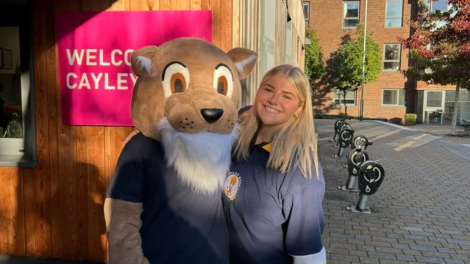 A person standing outside Cayley Hall posing with the hall’s mascot in a large animal costume, in front of a pink “Welcome to Cayley” sign on a sunny day.