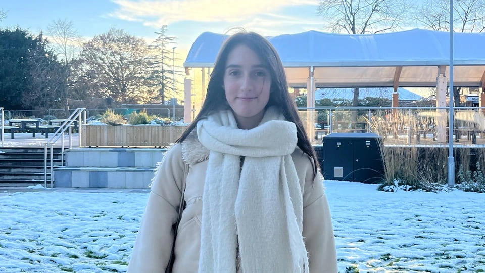 Student facing camera and smiling, she is wrapped up warm in winter coat and scarf. Behind her campus is covered in snow.