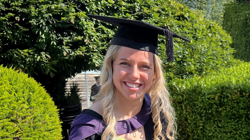 Student facing camera and smiling, she has curled blonde hair and is wearing a black and purple graduation gown, and black graduation cap.