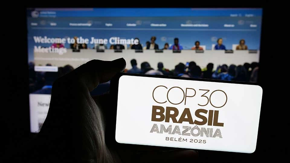 A person holding a smartphone displaying the logo for COP30 Brasil Amazônia Belém 2025. In the background, there is a blurred screen showing a climate meeting with the text “Welcome to the June Climate Meetings.”