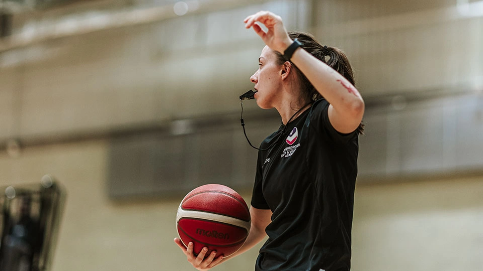 A woman with a whistle in her mouth, arm up, and basketball in her hand.