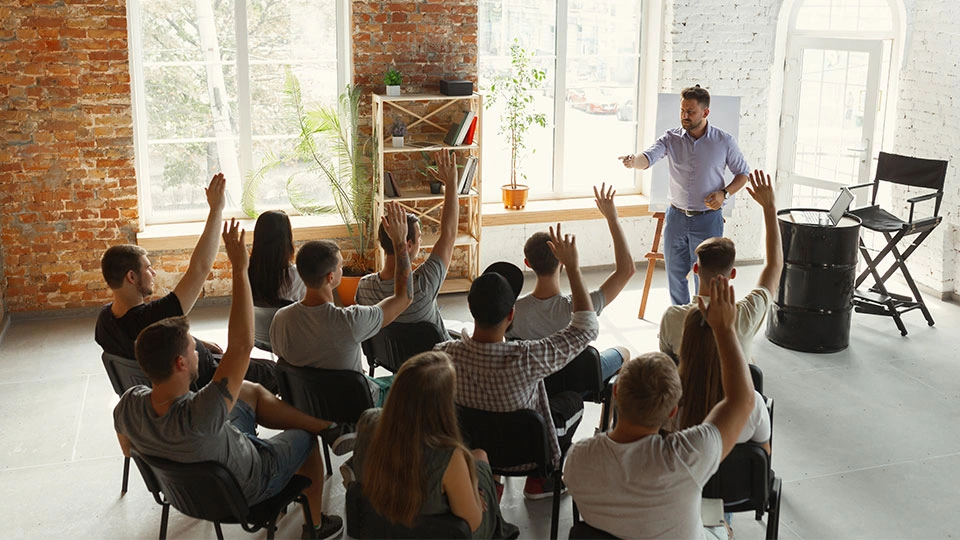 A group of people sat on chairs raising their hands opposite a lecturer who is standing and pointing towards them.