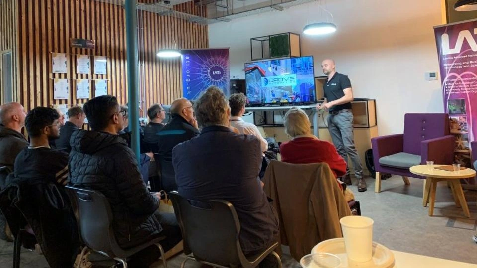 A man presenting to a seated audience with a tv screen behind him.