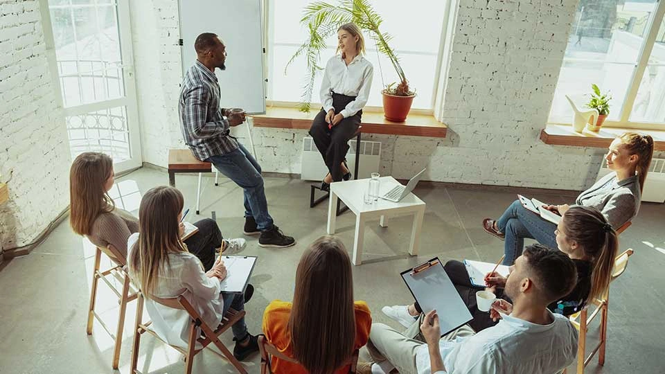 A group of people sat around a table talking, some are holding clipboards and pens.