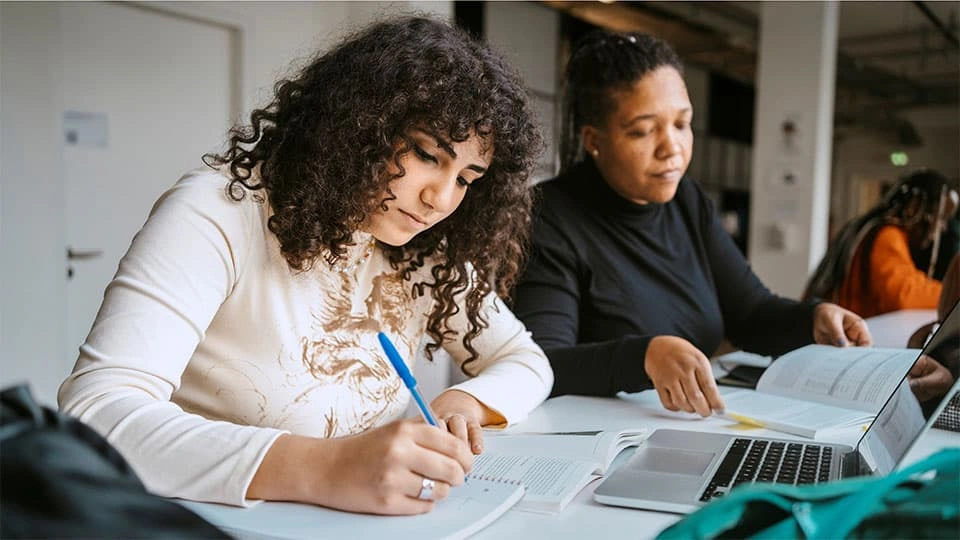 Student writing notes with a textbook and laptop in front of her in a library setting