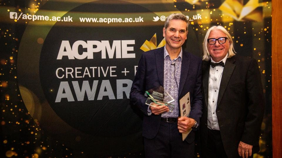 Two Loughborough Creative and Print Services team members holding a glass trophy standing in front of a black and gold awards backdrop reading 'ACPME CREATIVE + PRINT AWARDS'. 