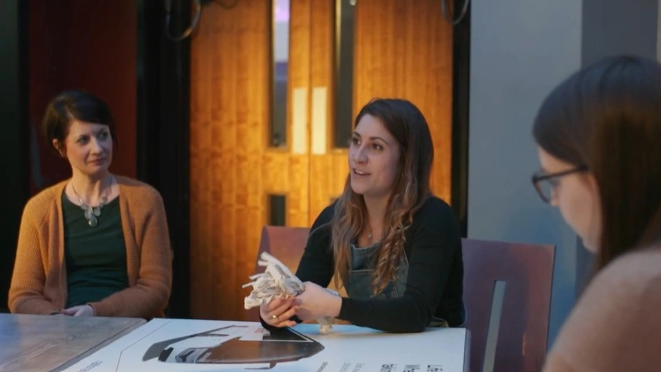Three people sitting at a table during an arts workshop, an arts worker is talking to the group holding macrame cord.
