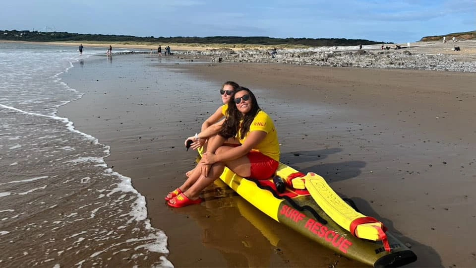 Beth Morgan smiling on a beach by the waterline in lifeguard gear