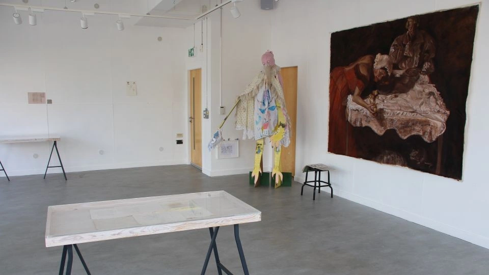 A gallery space with white walks and grey floor. A large unframed canvas painting is on the wall on the right with a birdlike textile sculptural piece hanging to the left of this and a display table in the foreground.