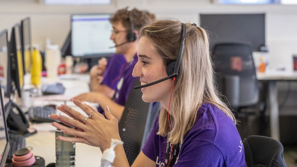 Close-up of two members of staff sat at computers, speaking into a headset.