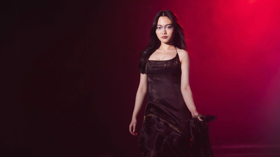 Model walking on a runway wearing a black dress with red lighting behind her.