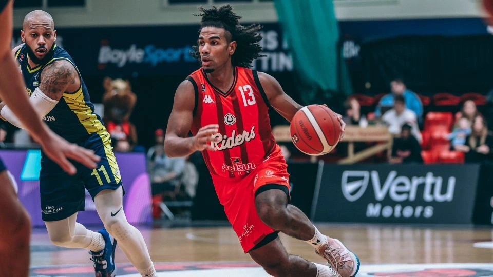 Connor playing mid-match on a basketball court with the ball, wearing the Red Leicester Riders kit and looking away from the camera. There is a player on the opposite team in the background 