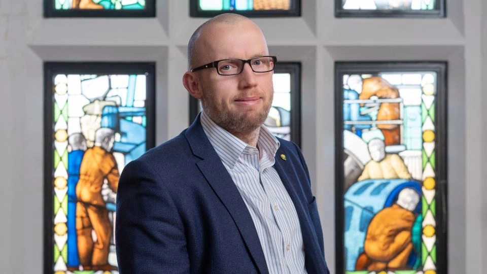 Photo of Martyn Edwards stood on his side with his face looking towards the camera. He is wearing a dark suit and is stood in front of stained glass windows inside the Hazlerigg Building