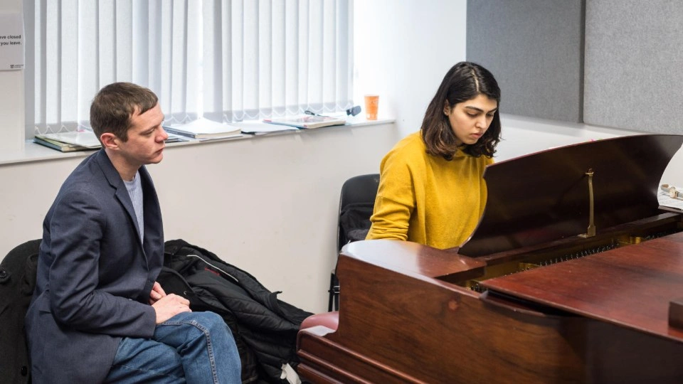 A piano teacher in a music room watching one of their students practice
