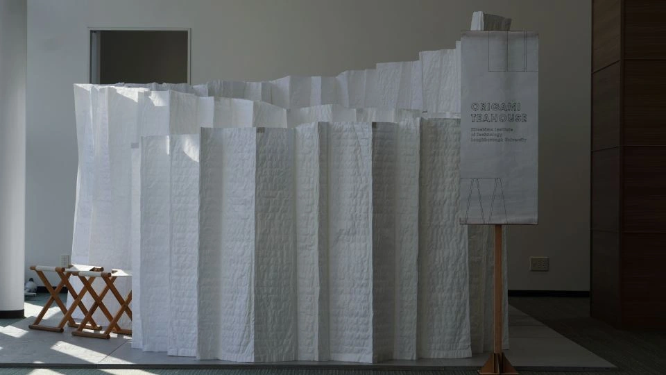 photo of the mobile origami teahouse designed by Loughborough students. The teahouse is made from white material, and has a banner in front of it, along with stools inside. 