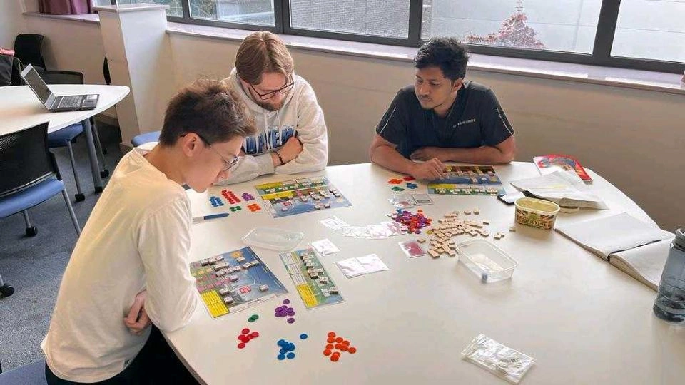 photo of three male students sat at a table working on a puzzle that has different coloured counters and various materials 