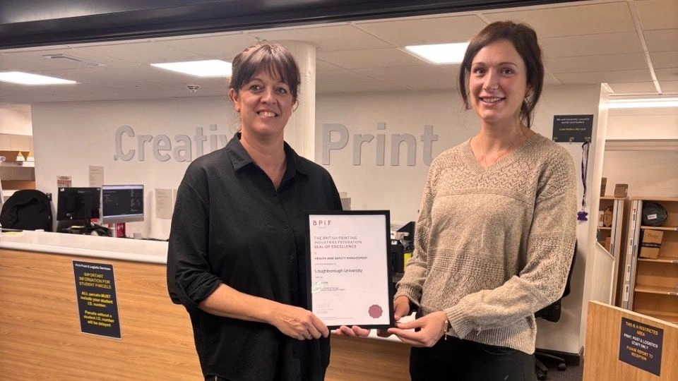 Two members of staff standing in front of a desk holding a certificate.