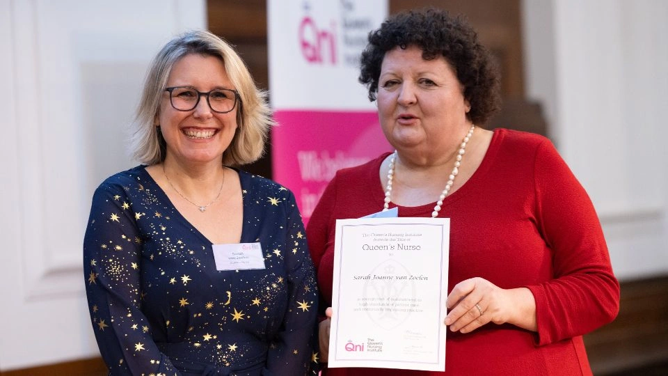 Two people standing together, the person on the right is holding a Queen’s Nurse certificate.
