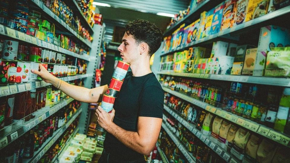 A student holding a stack of tins in the aisle of a supermarket.