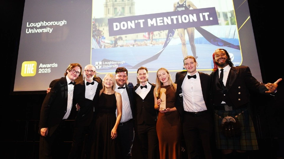 A group of people in formal attire stand on stage holding an award, with a large screen behind them showing 'DON’T MENTION IT,' the Loughborough University logo, and 'THE Awards 2025.'